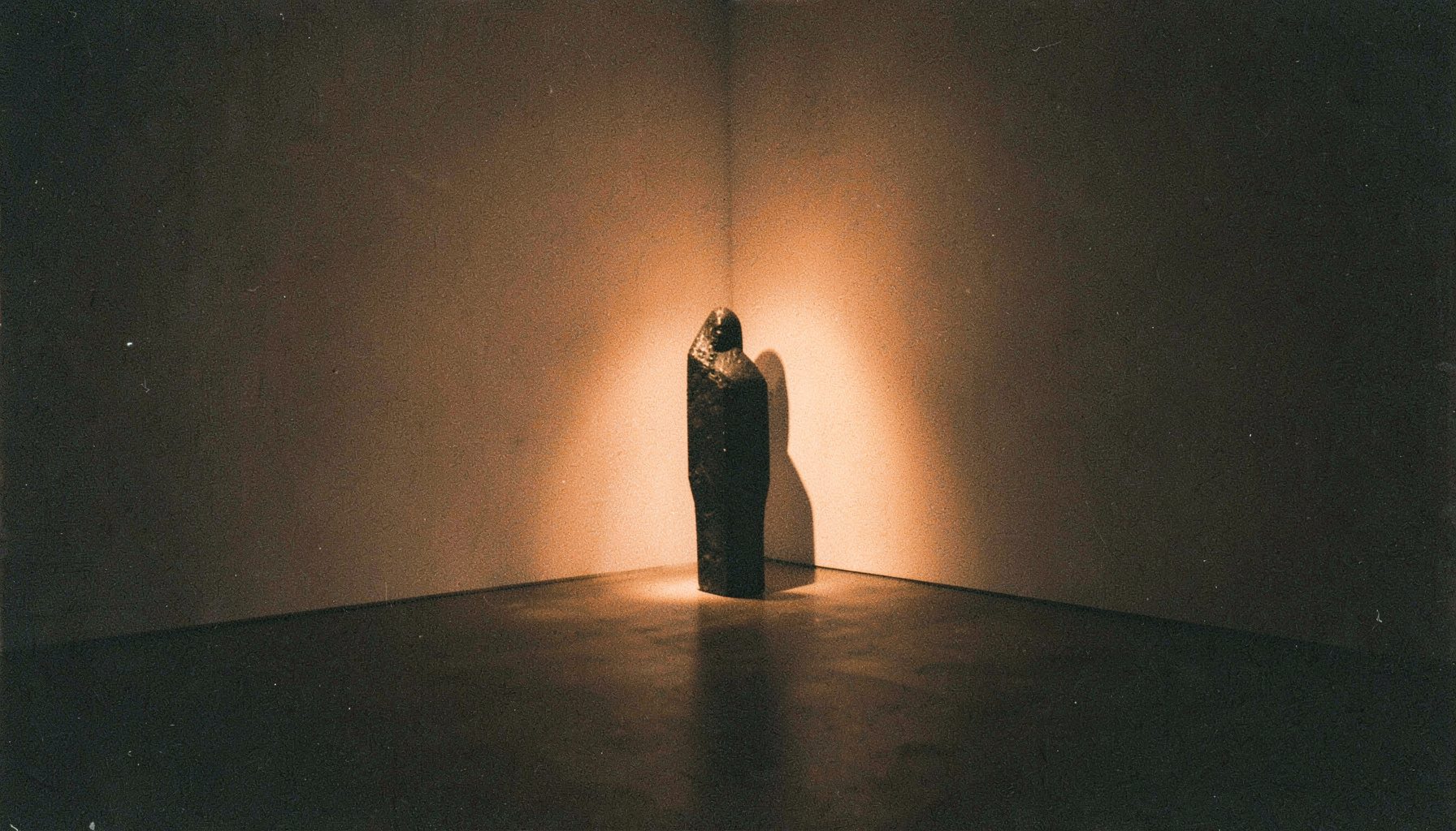 Fotografía analógica granulada de un espacio expositivo minimalista con una escultura abstracta iluminada por un haz de luz cálida, tonos carbón y ámbar, atmósfera íntima y editorial.