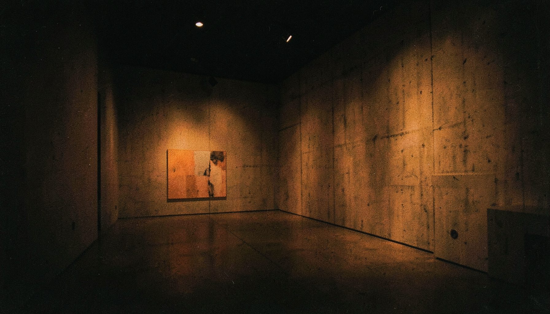 Una fotografía analógica con grano visible de una galería de arte contemporáneo en penumbra, dominada por negros profundos y acentos de luz ámbar dorada sobre paredes de hormigón, con un ambiente sereno y cinematográfico.